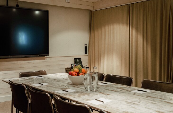 Mötesrum med stor TV, marmorbord, stolar, fruktskål och vattenflaskor. Beige gardiner och synliga träpaneler. Hesselby Slott, Hässelby